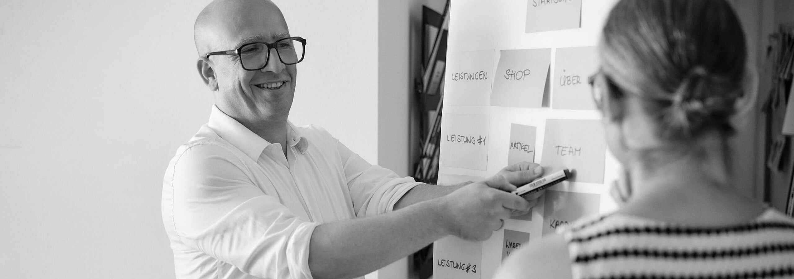Designer zeigt auf Flipchart mit Post-Its mit einer Websitstruktur, Kundin schaut auf das Flipchart